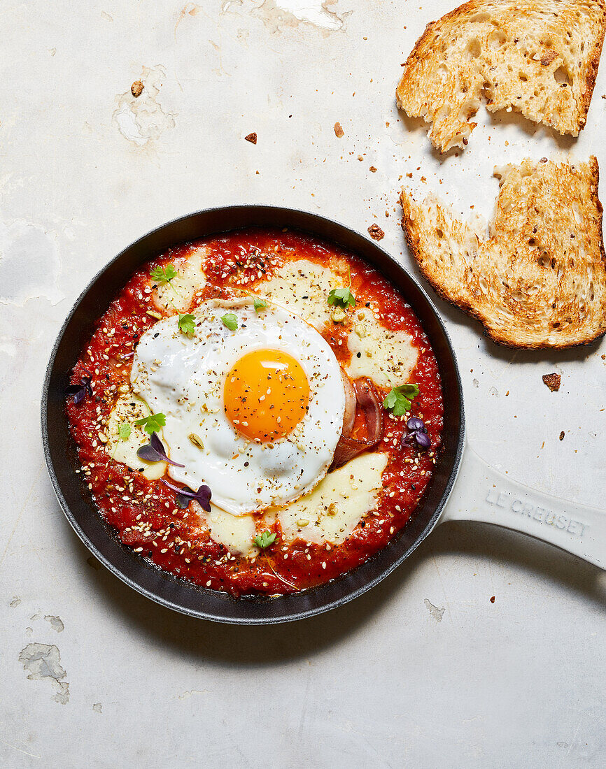 Top view of frying pan with appetizing shakshuka with egg and cheese placed near bread on table