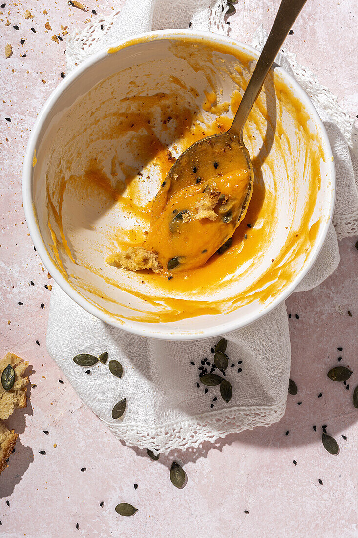 Draufsicht auf einen Löffel in einer weißen Keramikschüssel mit gegessener Kürbiscremesuppe, die auf dem Tisch neben verstreuten Kernen und knusprigen Brotstücken steht