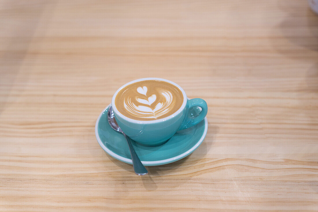 Becher mit heißem Kaffee mit kreativer Latte Art, serviert auf einem Teller mit Teelöffel auf einem Holztisch in einem modernen hellen Kaffeehaus