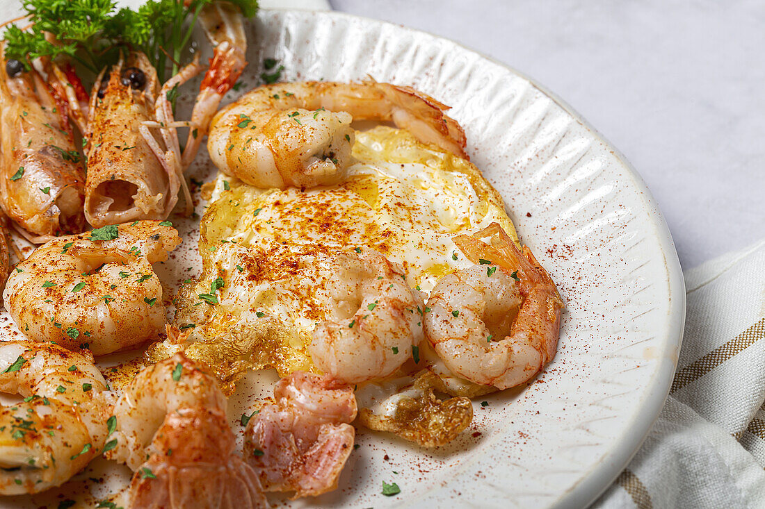 From above appetizing fried eggs with king prawns served on white plate with green herbs and seasoning on towel in light kitchen