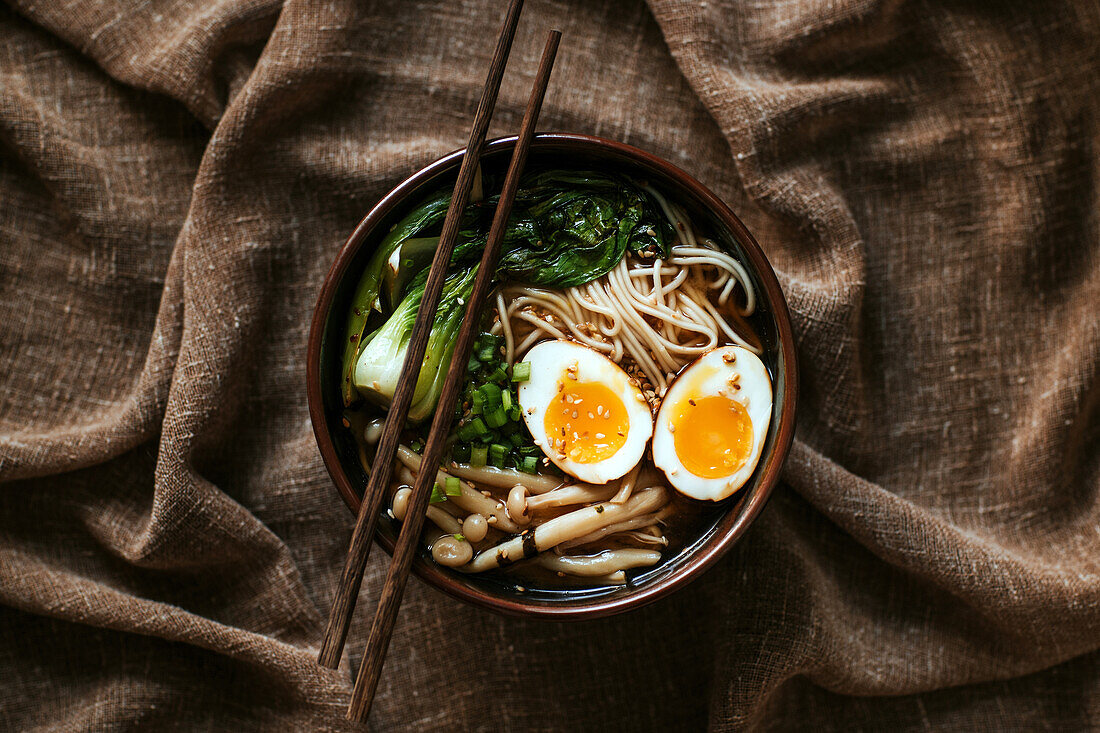 Top view of ceramic bowl with delicious ramen and chopsticks placed on table covered with brown tablecloth