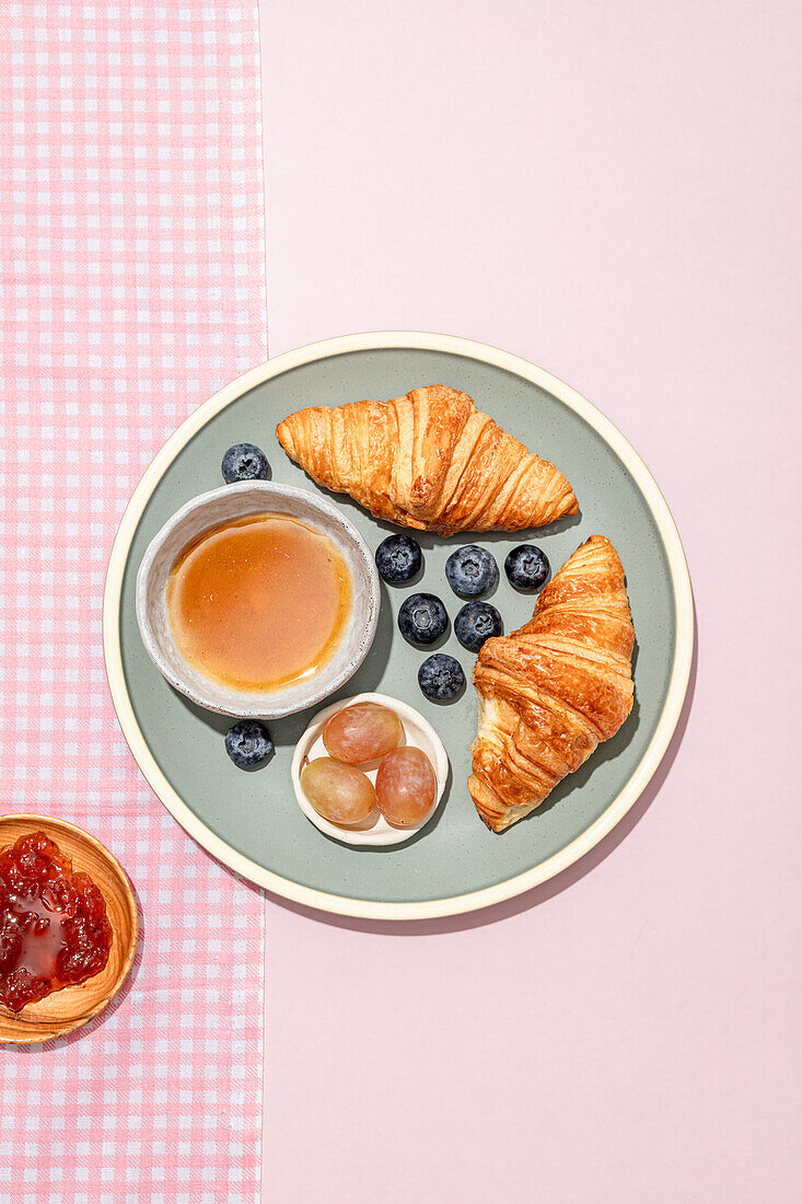 Draufsicht auf leckere Croissants auf Keramikteller mit frischen Blaubeeren und Marmelade auf dem rosa Tisch