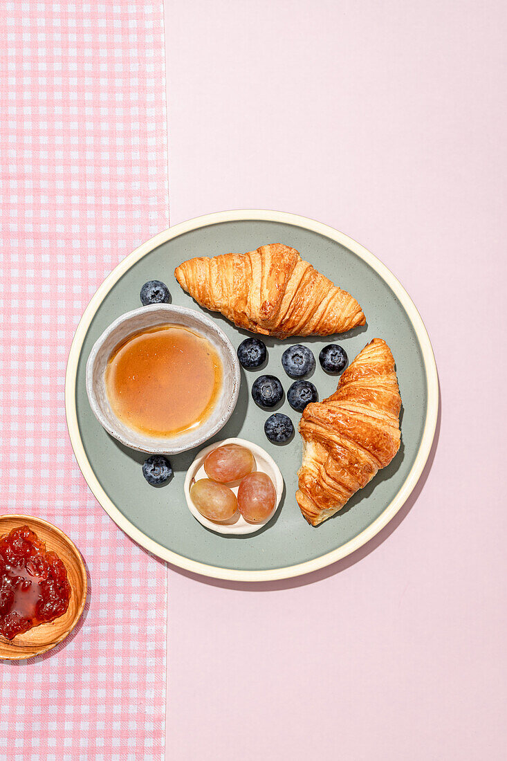 Top view of delicious croissants served on ceramic plate with fresh blueberries and jam placed on pink table