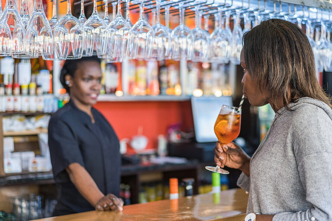 African American female customer drinking cold fresh cocktail with straw in bar and looking at bartender