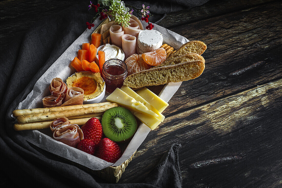 Von oben Brunch-Box mit verschiedenen Wurst- und Käsesorten und Knäckebrot in der Nähe von reifen Kiwis, süßen Erdbeeren und geschälten Mandarinen in der Nähe von Marmelade in einem Glas auf einem Holztisch in der Nähe einer Serviette