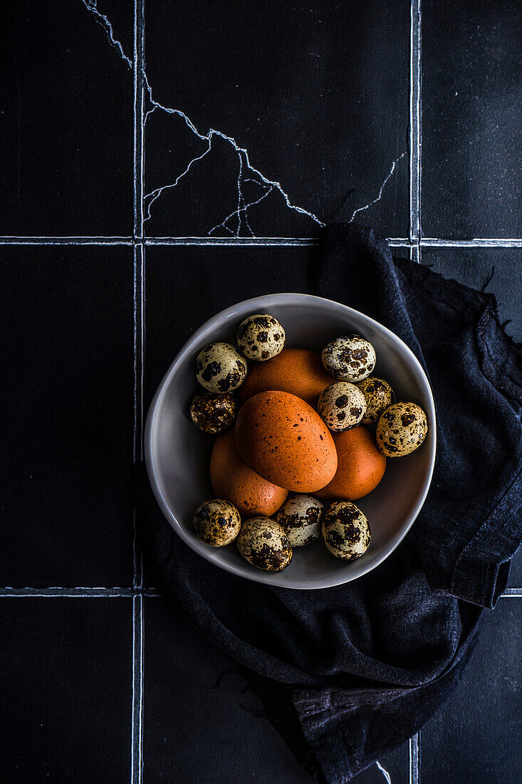 From above ceramic bowl with raw quail and chicken eggs on tiled table