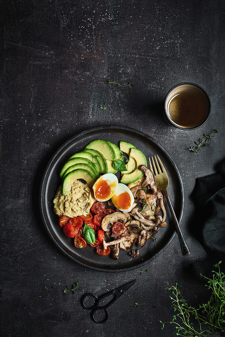 Überkopf gefüllter Teller mit köstlichem Hummus aus gebratenen Champignons und gekochten Eiern mit Tomaten und Avocado, serviert mit frischen Basilikumblättern