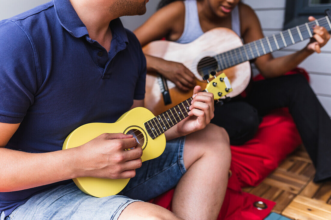 Unkenntlich gemachtes multiethnisches Paar sitzt auf Sitzsäcken auf der gemütlichen, hellen Terrasse einer Wohnung und spielt Gitarre und Ukulele