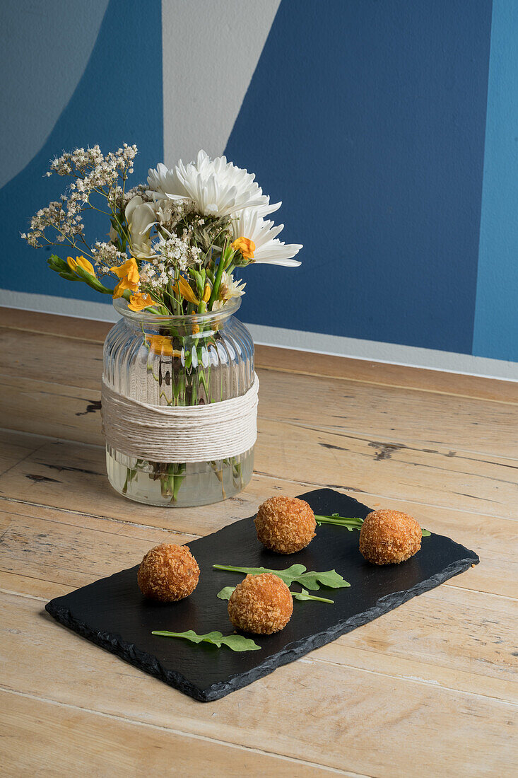 Blick von oben auf appetitliche Käsebällchen, die auf einem schwarzen Brett neben einer transparenten Vase mit blühenden Blumen serviert werden