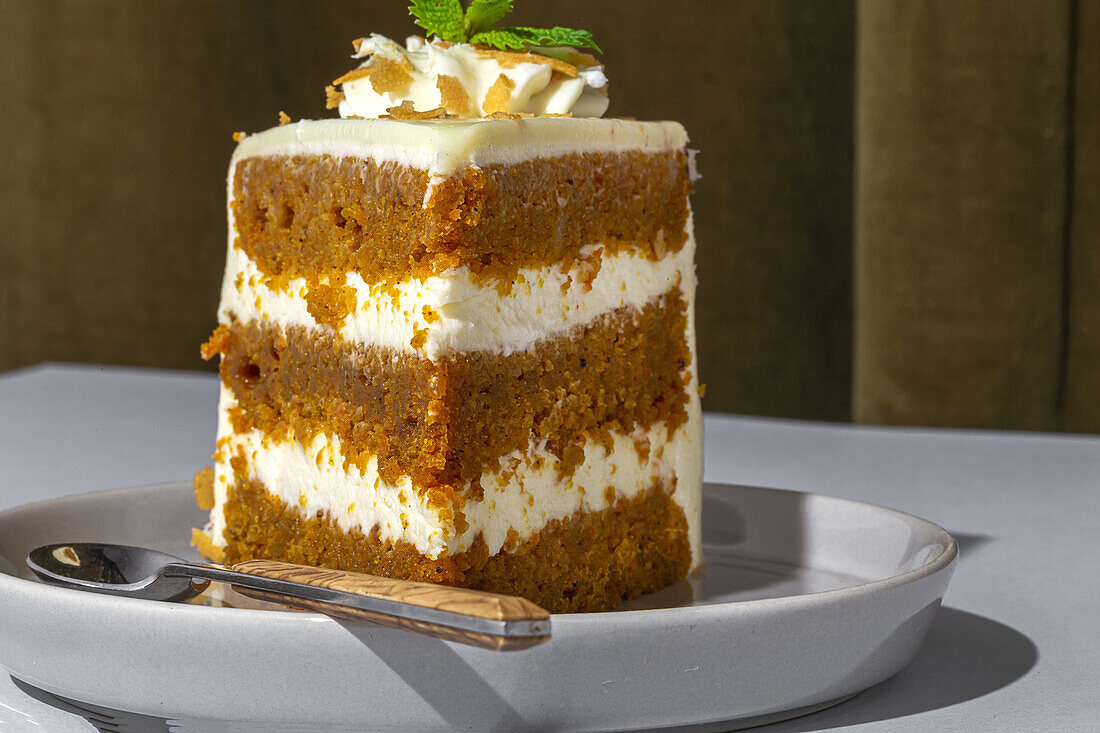 Slice of tasty sweet carrot sponge cake with cream decorated with mint leaf served on plate with spoon on table in light room with curtain on the background