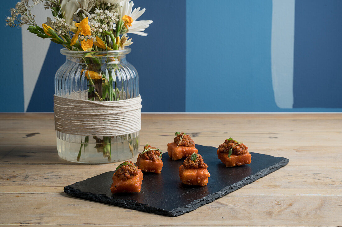 Von oben köstliche Vorspeise in quadratischen Bissen serviert auf schwarzem Brett in der Nähe Strauß blühender Blumen in Glasvase gegen blaue Wand