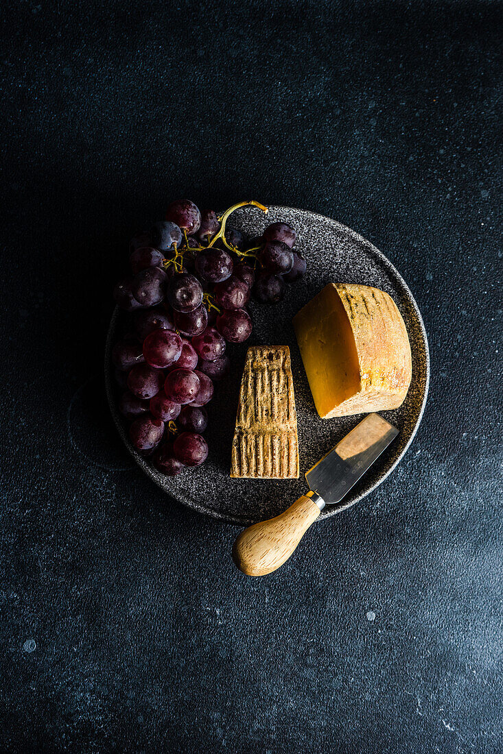 Von oben schwarzer Steinteller mit Käse und rohen, schmackhaften Trauben auf dunklem Betontisch