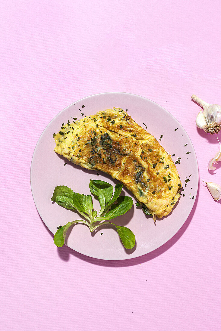 Leckeres Omelett auf einem Teller mit frischen Petersilienzweigen und Knoblauchzehen auf rosa Hintergrund