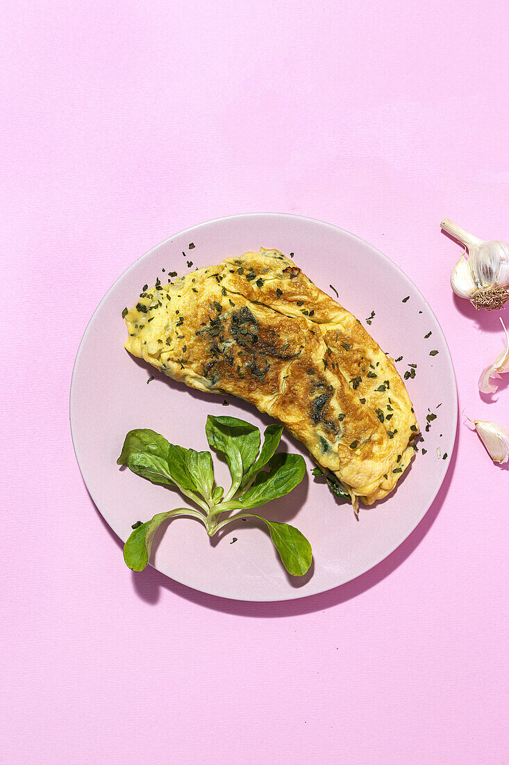 Tasty omelette on plate against fresh parsley sprigs with garlic cloves on pink background