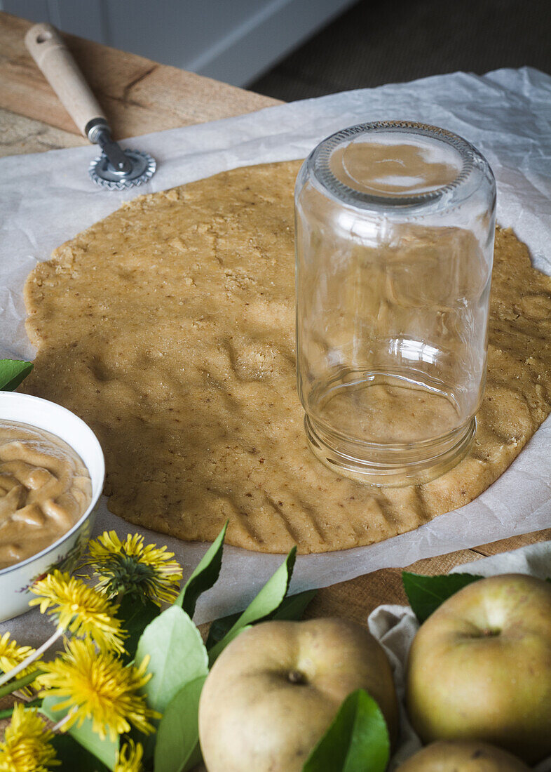 From above glass jar placed on thin fresh dough near apples and dandelions during pastry preparation in kitchen at home