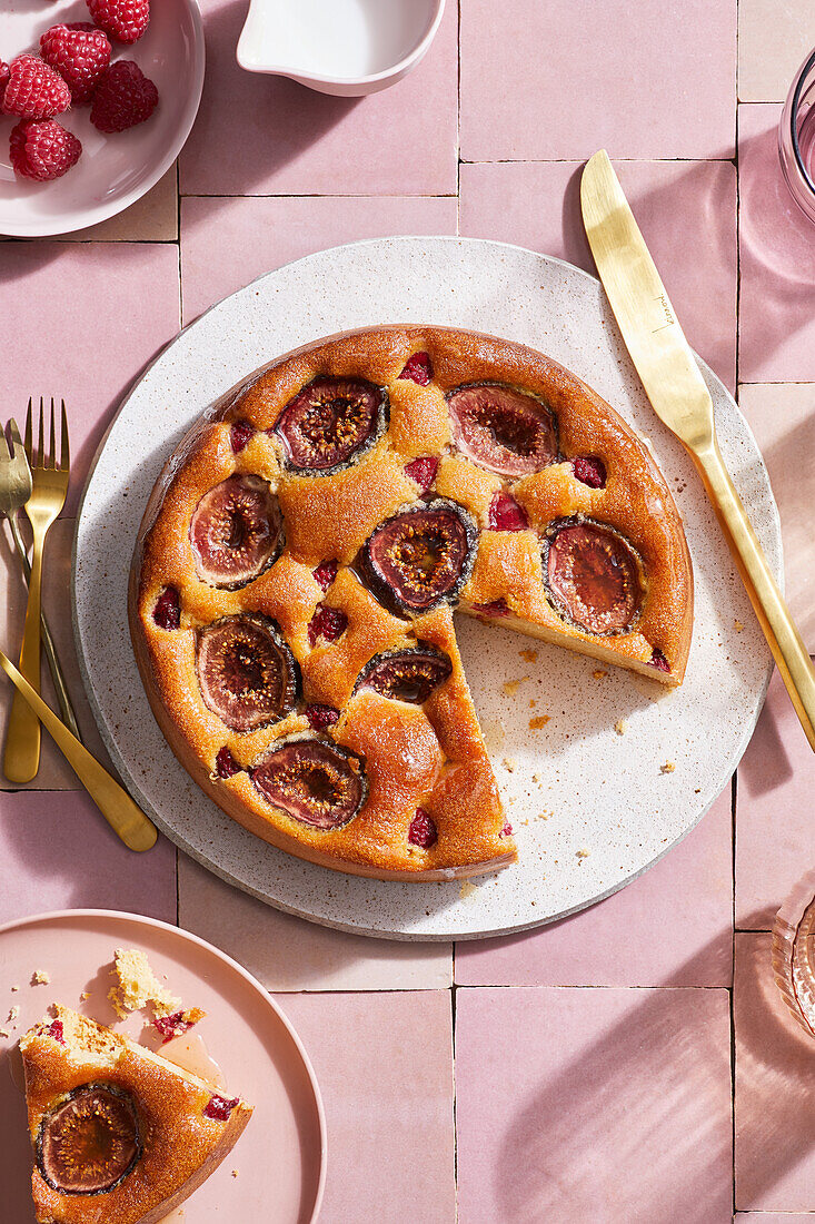 Draufsicht auf appetitlichen hausgemachten spanischen Kuchen mit Feigen, der auf einem runden Teller neben goldenem Besteck auf einer rosa Fliese serviert wird