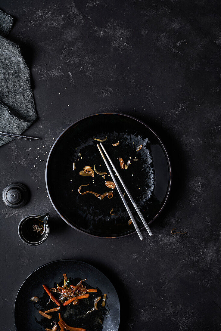 Overhead black empty bowl with pair of chopsticks and leftovers of Korean cuisine Japchae at table near full plate