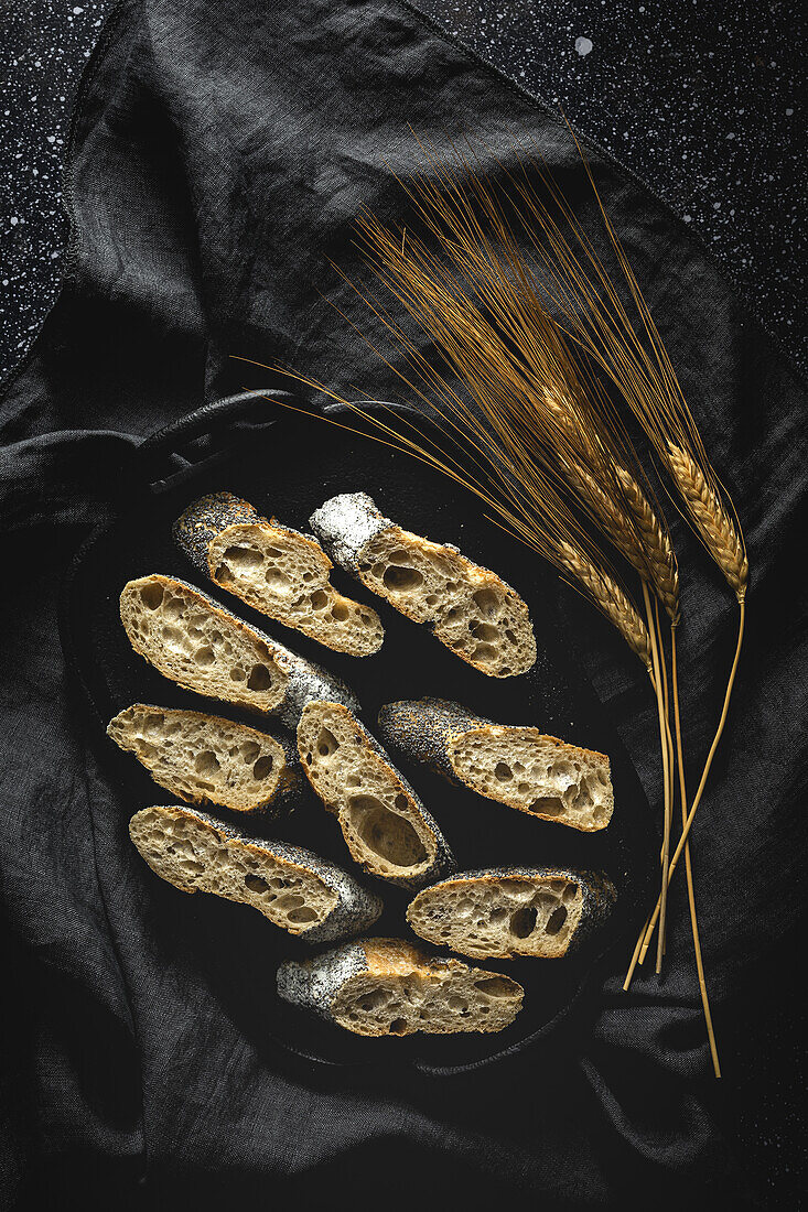 From above of appetizing crusty bread near wheat spikes and dark fabric on table