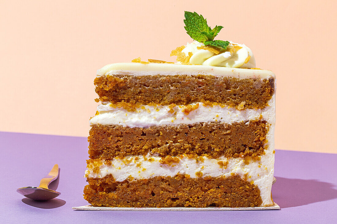 Close up of slice of tasty sweet carrot sponge cake with cream decorated with mint leaf served on plate with spoon on table on colorful background