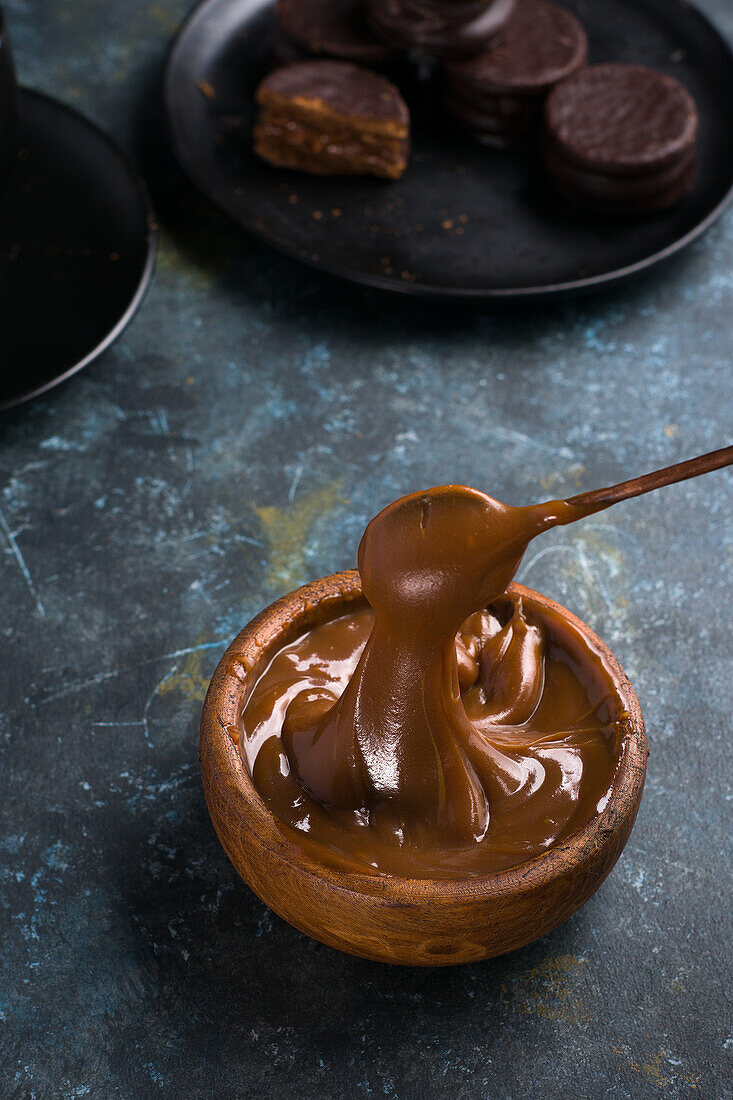 Dulce de leche in brown bowl against black cup and saucer near chocolate alfajores