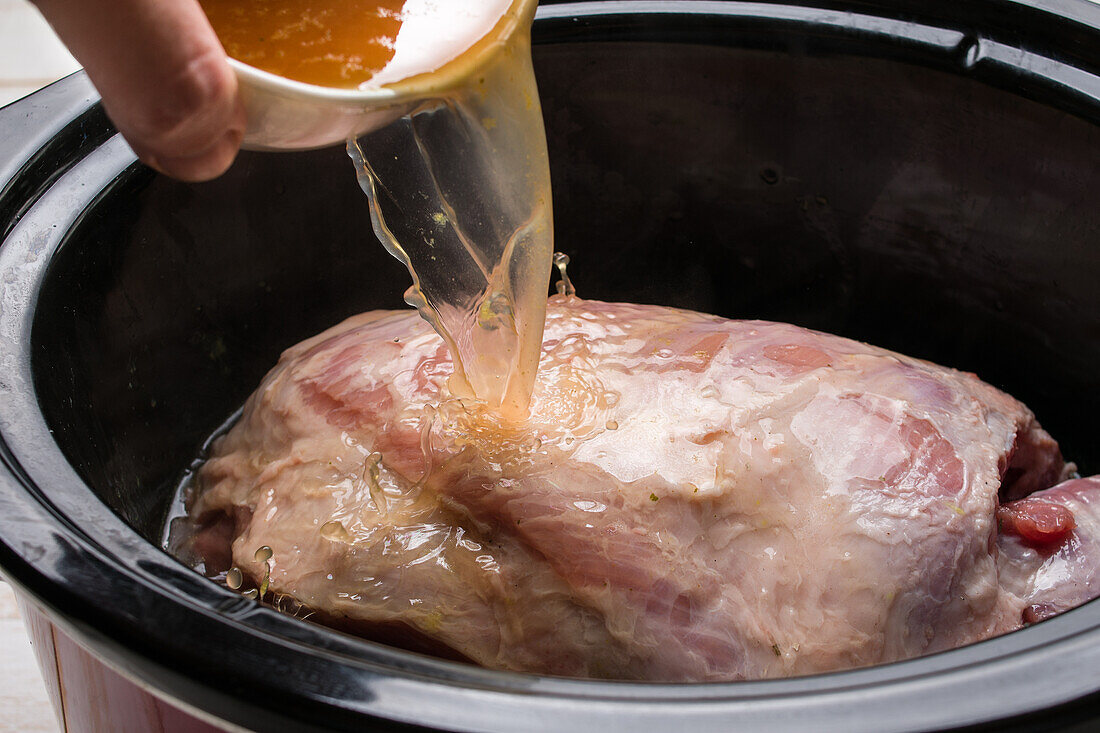 Anonyme Person gießt aromatische Brühe über ein Stück rohes Fleisch im langsamen Kocher, während sie eine kubanische Schüssel zubereitet