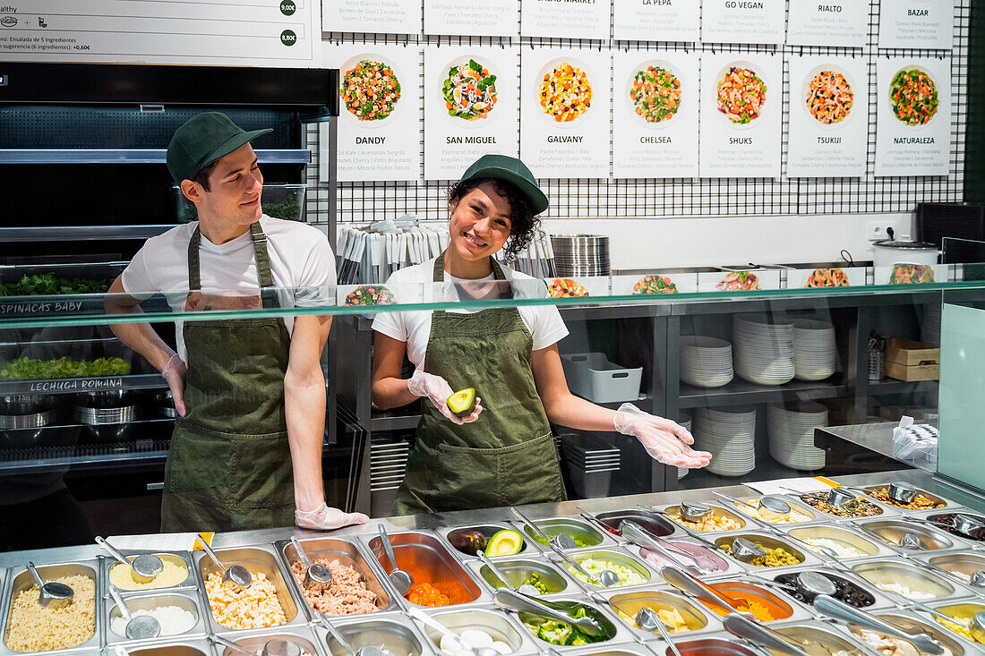 Zufriedene multiethnische Verkäuferinnen und Verkäufer in Uniform und Handschuhen stehen an der Theke mit verschiedenen Salatzutaten im Supermarkt