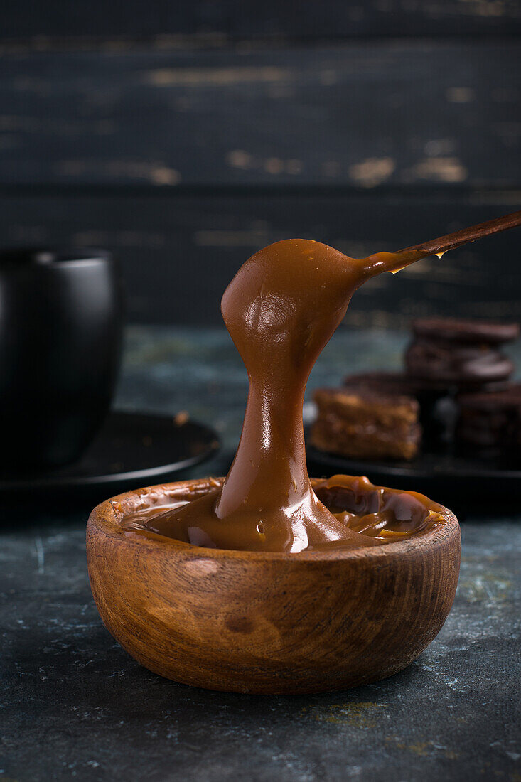 Dulce de leche in brauner Schale vor schwarzer Tasse und Untertasse neben Schokoladen-Alfajores