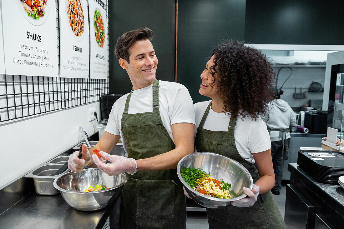 Positive multiracial male and female sellers in aprons preparing healthy salads while working in supermarket bar and looking at each other
