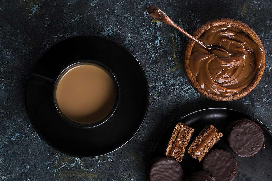 Draufsicht auf Schokoladen-Alfajores auf einem Teller und Dulce de Leche in einer Schale neben einer Tasse mit heißer Schokolade