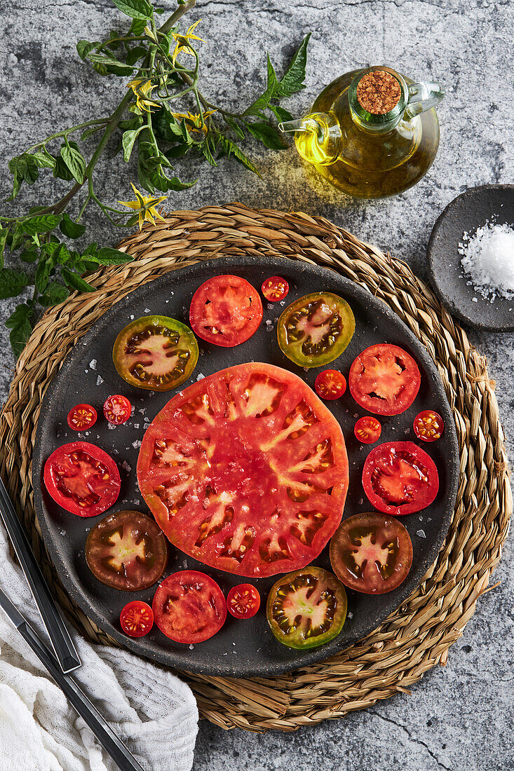 Top view of delicious sliced tomatoes in cast iron plate placed on wicker napkin near plate with sea salt and jug of olive oil on concrete table