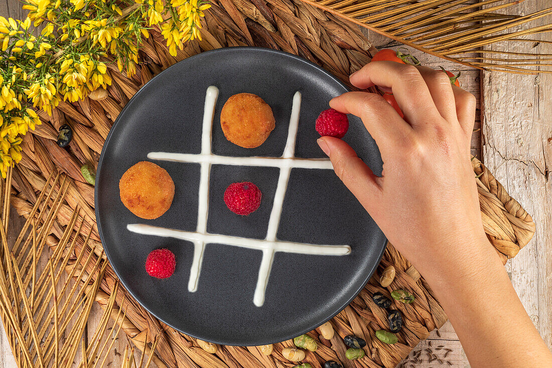 Top view of crop unrecognizable cook putting whole raspberry on plate with fritters and whipped cream representing noughts and crosses game