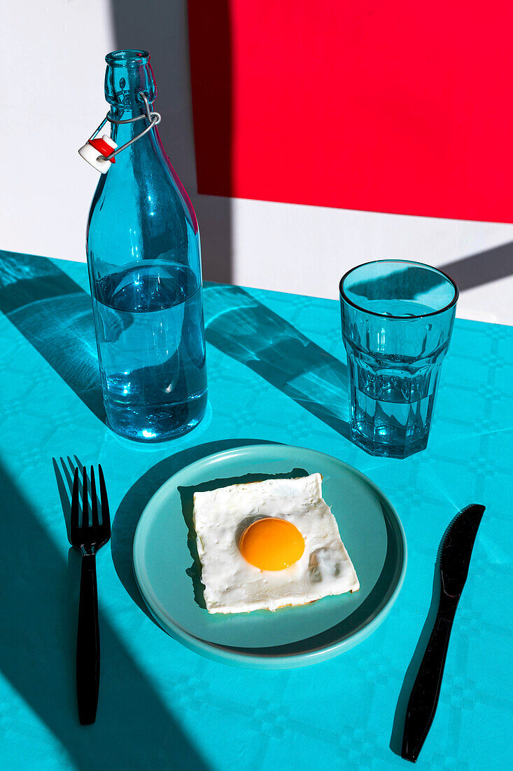 Von oben Komposition von serviertem frischem Spiegelei auf Teller mit Messer und Gabel gegenüber Flasche Wasser und Glas auf blauem Tisch vor rotem Hintergrund im Sonnenlicht