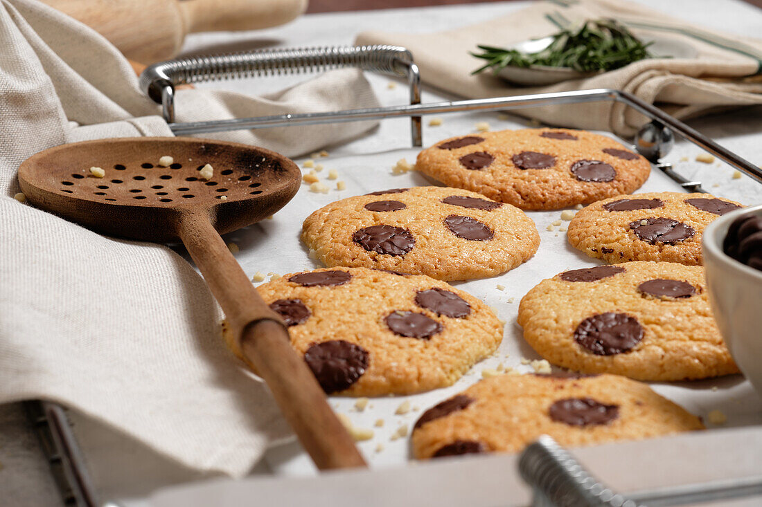 Blick von oben auf frisch gebackene süße Kekse mit Schokoladensplittern auf einem Metallgitter auf einem Tisch mit verschiedenen Küchengeräten und grünen Rosmarinzweigen