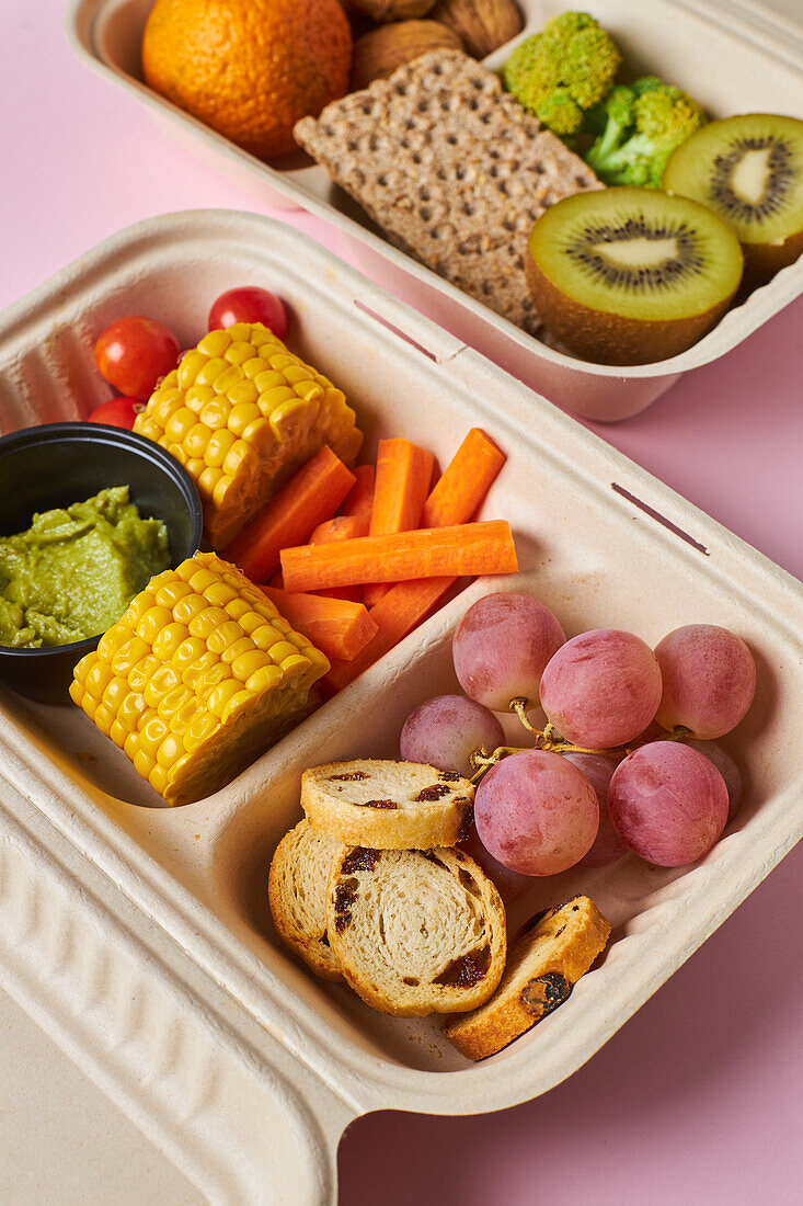 From above of lunch boxes with healthy food including crackers carrot sticks grapes cherry tomatoes with kiwi broccoli walnut and tangerine on pink background