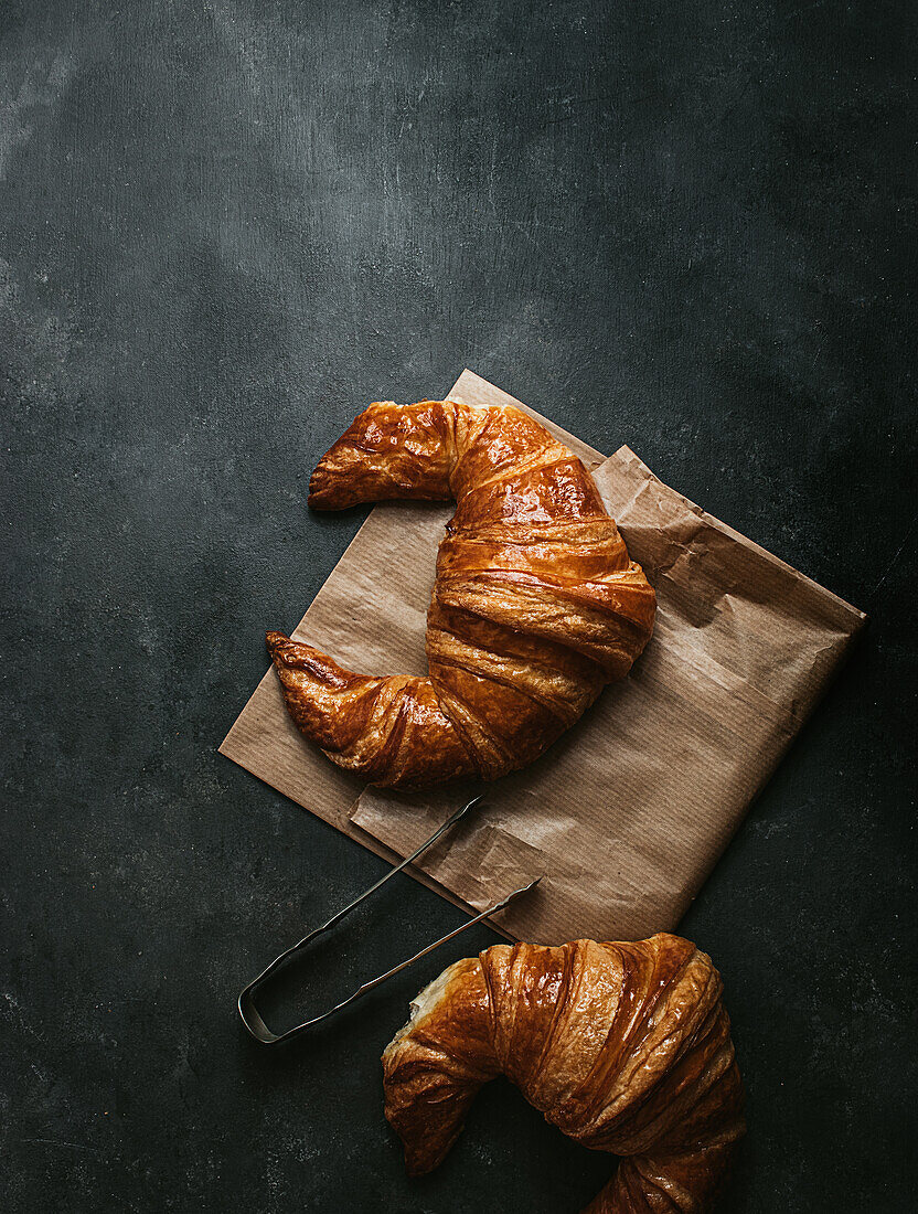 Top view Zusammensetzung mit leckeren frischen knusprigen Croissants mit Papiertüte und Metallzange auf schwarzem Hintergrund platziert