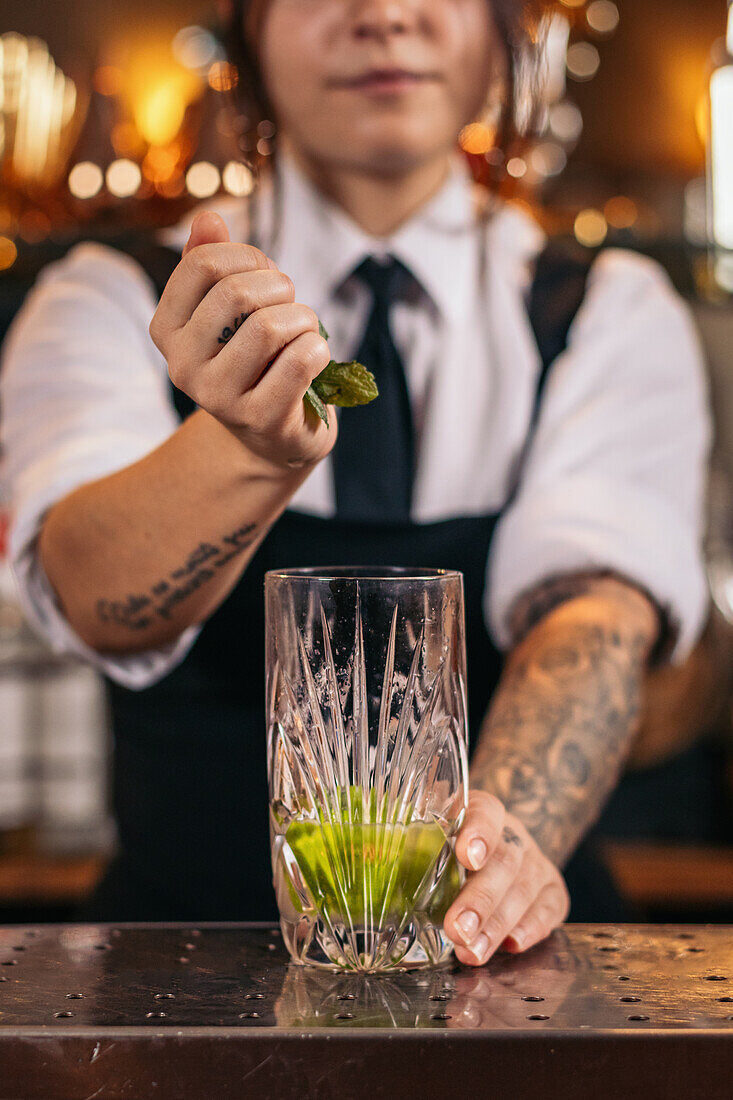 Crop Barkeeper, der frische Minzblätter in ein Glas gibt, während er einen Mojito-Cocktail am Tresen einer Bar zubereitet