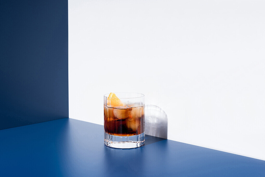 Glass of alcoholic cocktail made with red vermouth served with orange slices in light studio with blue and white wall