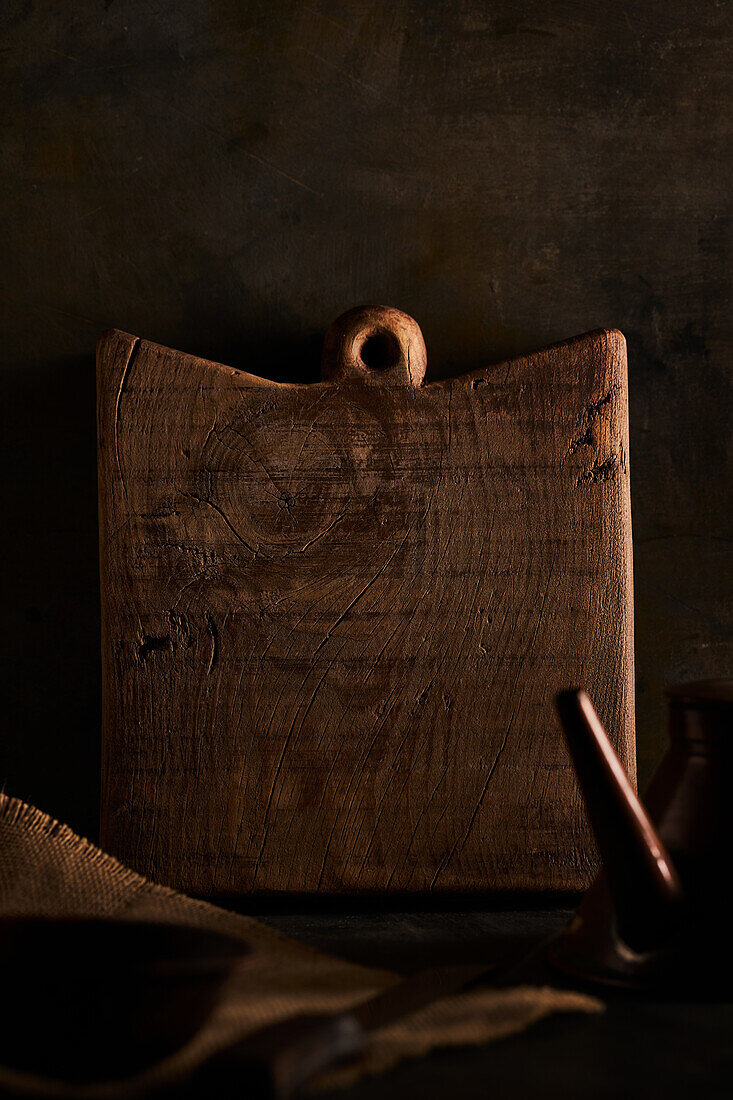 Braunes Hartholzschneidebrett in Form eines Quadrats auf einem Tisch mit Messer und Schüssel in einer dunklen, rustikalen Küche