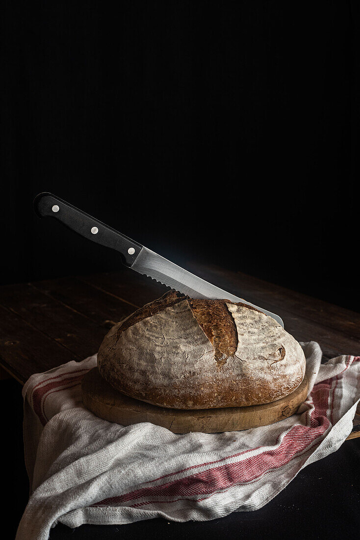 Hausgemachtes Sauerteigbrot mit knuspriger Kruste und Brotmesser auf rustikalem Holzbrett auf schwarzem Hintergrund