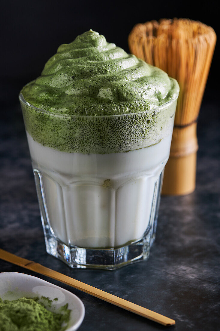 Glass of fresh dalgona coffee with matcha and Japanese tea ceremony accessories consisting of bamboo whisk called chasen and traditional scoop named chashaku placed on gray surface