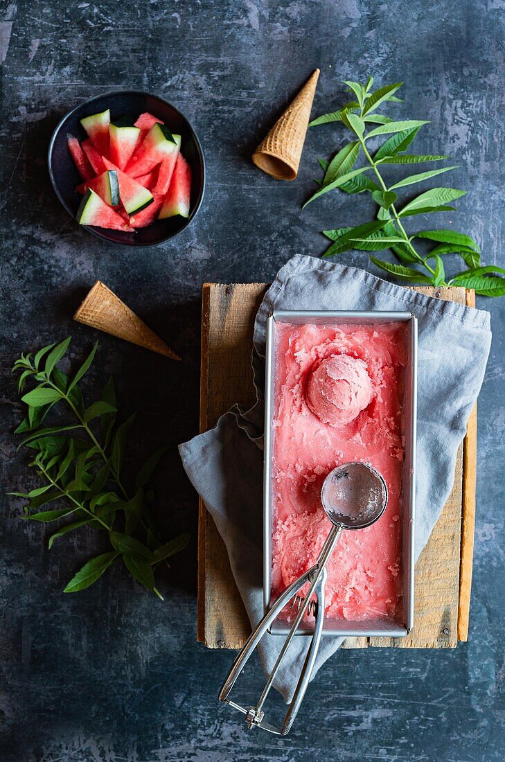 Top view of container with sweet watermelon sorbet and scoop placed on napkin and board near mint twigs and cornets on gray table