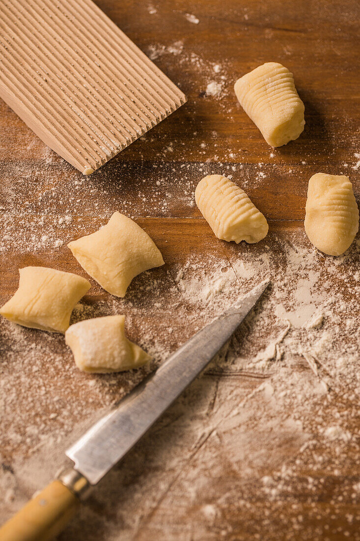 Top view of pieces of soft raw dough placed on wooden table covered with flour near ribber board and knife during gnocchi preparation in the kitchen