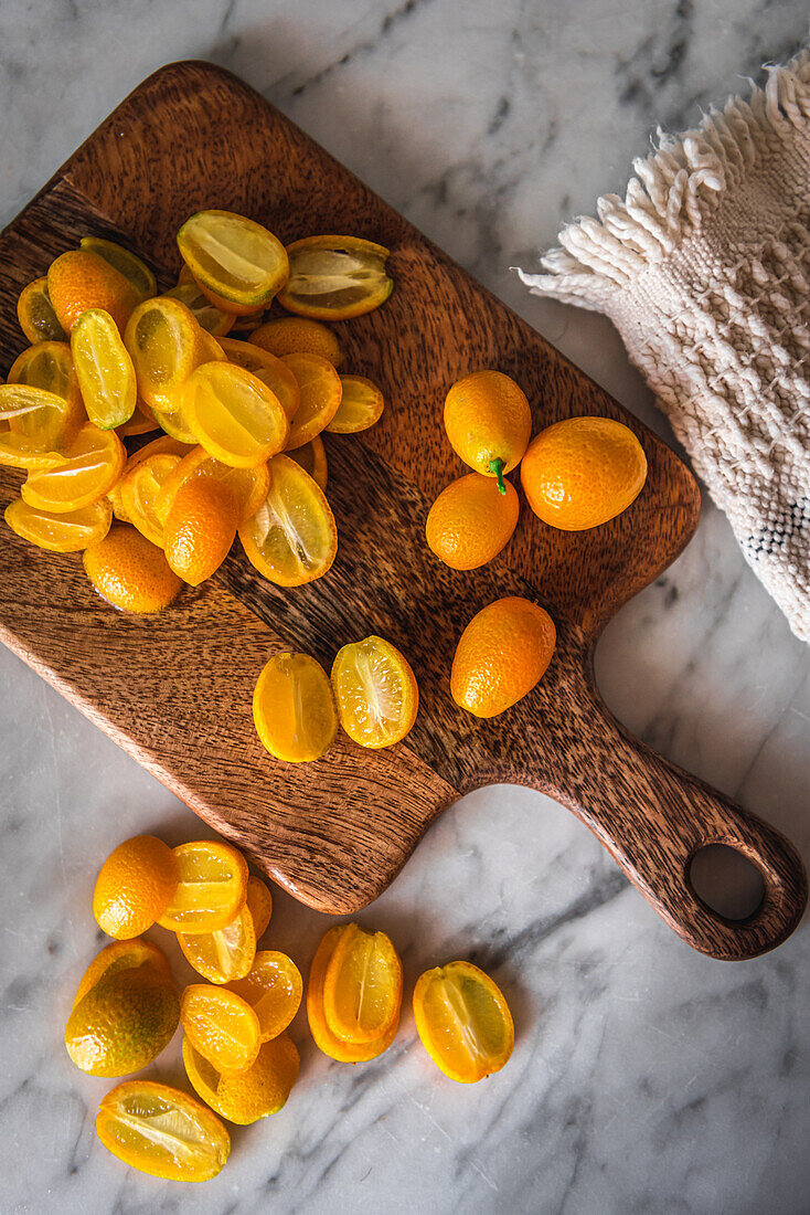 Draufsicht auf einen Stapel frischer, in Orangen geschnittener Kumquats auf einem hölzernen Schneidebrett auf einem Marmortisch mit Handtuch in der Küche