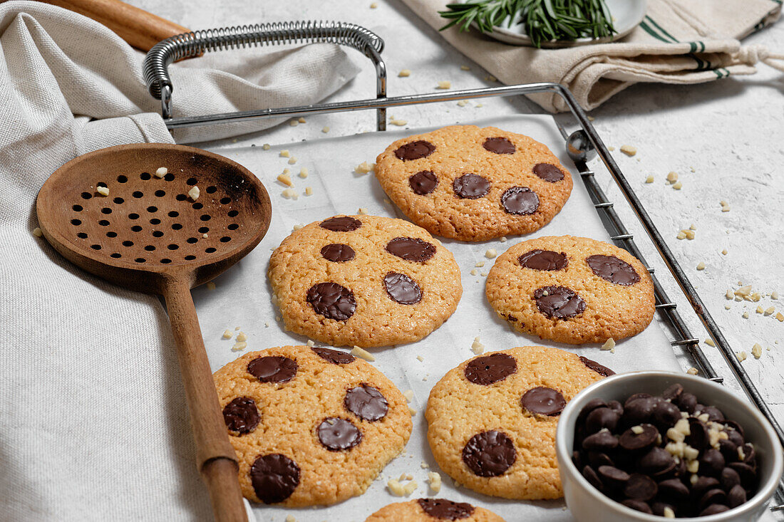 From above of freshly baked sweet cookies with chocolate chips on metal grid placed on table with various kitchen tools and green rosemary branches