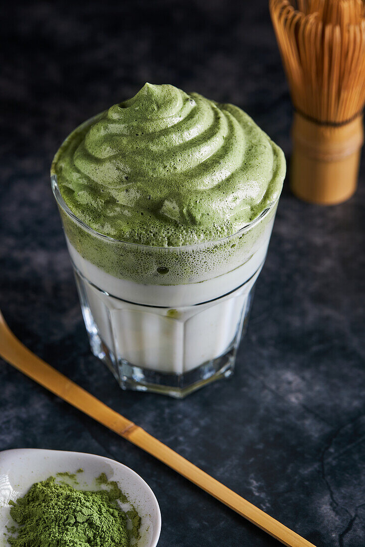 From above of glass of fresh dalgona coffee with matcha and Japanese tea ceremony accessories consisting of bamboo whisk called chasen and traditional scoop named chashaku placed on gray surface