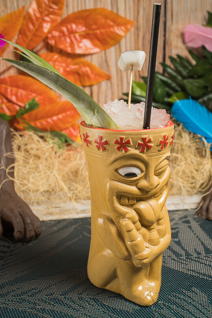 Polynesischer Tiki-Becher mit kaltem alkoholischem Getränk, dekoriert mit Stroh und grünen Blättern vor bunten Blättern und trockenem Gras