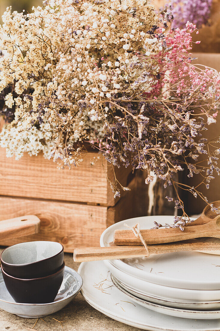 Schwarzes und weißes Keramikgeschirr mit Holzlöffeln neben einer braunen Eichenkiste mit einem üppigen Strauß bunter Wildblumen auf einem Tisch im Herbstgarten