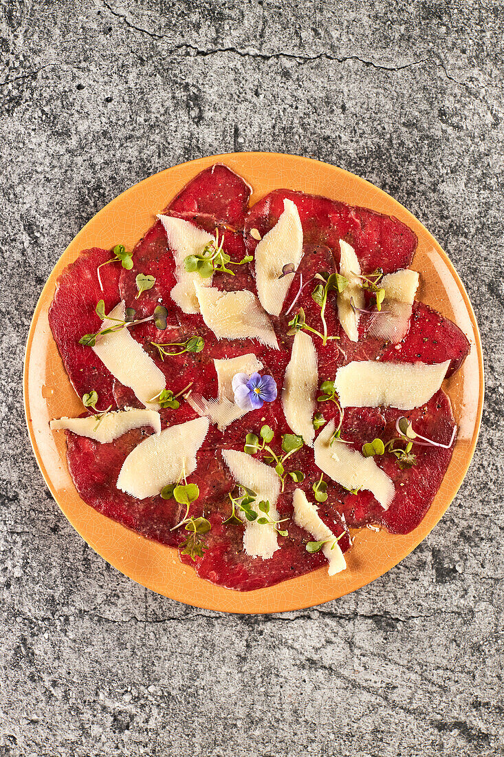 From above slices of veal with lemon and oil with black pepper garnished with sprouts and slices of parmesan cheese and served on plate on gray marble table in restaurant