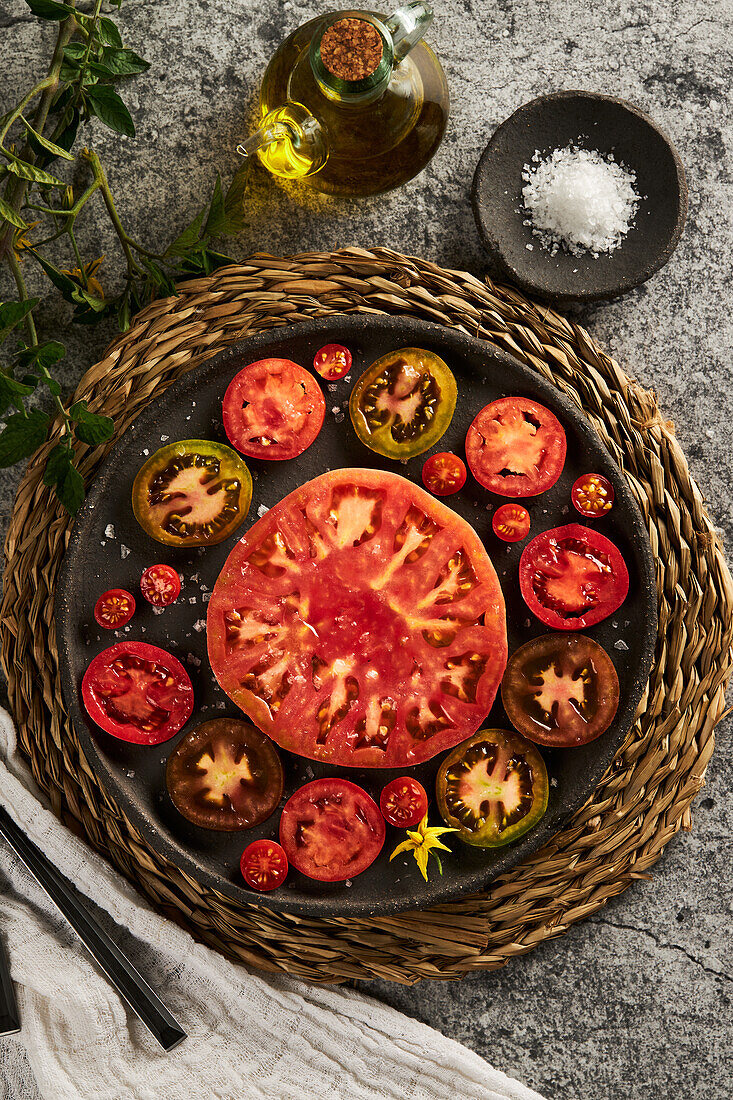 Top view of delicious sliced tomatoes in cast iron plate placed on wicker napkin near plate with sea salt and jug of olive oil on concrete table