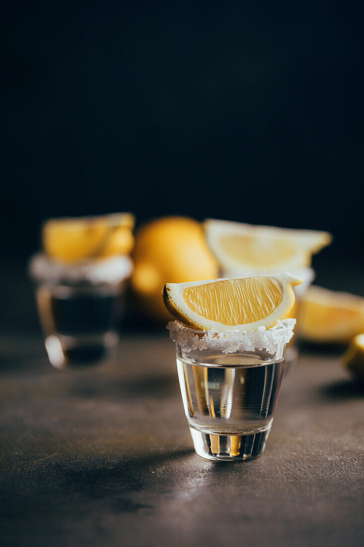 Tequila-Shots mit Salz und Zitrone auf spiegelnder Oberfläche vor dunklem Hintergrund